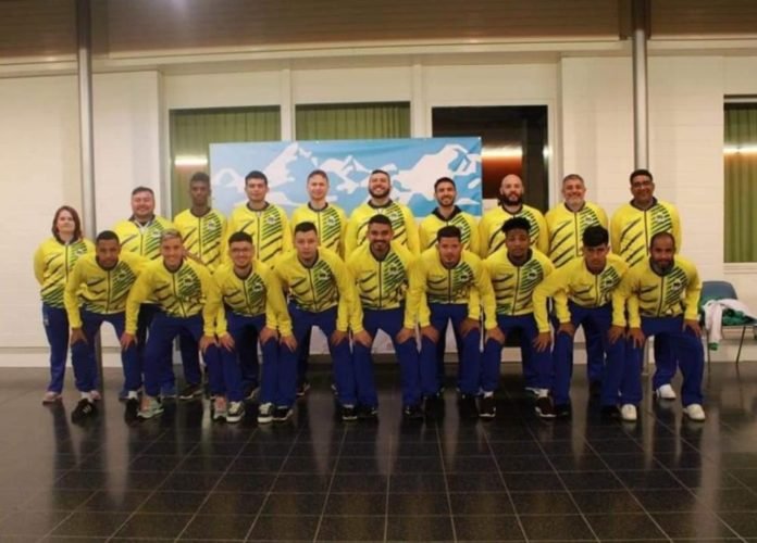#descriçãodaimagem: Foto dos atletas da delegação da Seleção Brasileira de Futsal de Surdos. Eles estão enfileirados em dois grupos. Um grupo atrás, em pé, e outro grupo na frente, abaixado. Todos usam o uniforme com blusão amarelo com detalhes verdes e calça azul marinho. Fim da descrição.
