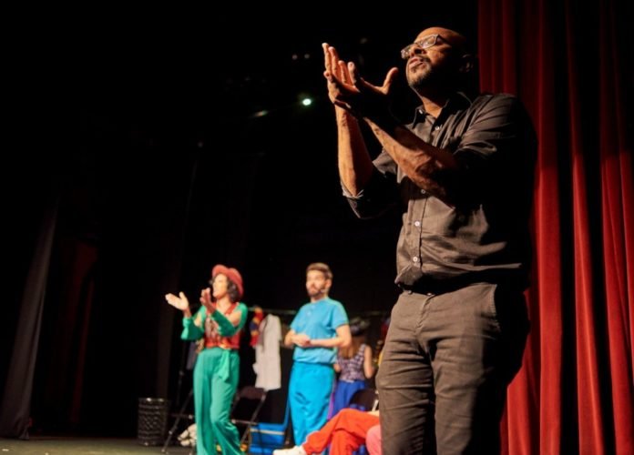 #descriçãodaimagem: Foto de uma apresentação teatral com interpretação em Libras. Ao fundo vemos os atores no palco e em primeiro plano, vemos o intérprete de Libras, traduzindo a apresentação. Fim da descrição.