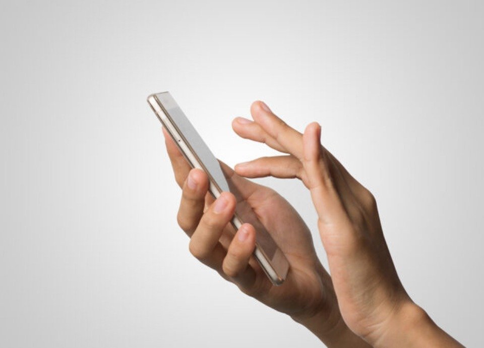 #descriçãodaimagem: Foto de uma mão segurando o celular e a outra mão está teclando na tela do aparelho. Fim da descrição.