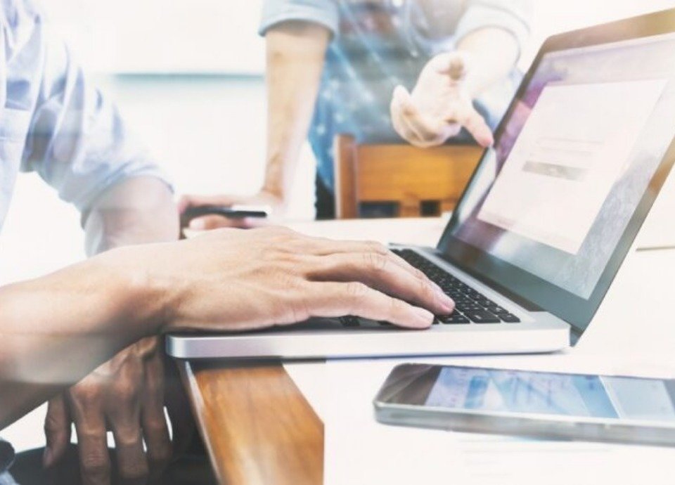 #descriçãodaimagem: Foto de uma mesa de trabalho. A mão de uma pessoa está sobre o teclado de um notebook. Um celular está sobre a mesa, ao lado do computador. Ao fundo uma outra pessoa está apoiada na mesa. Fim da descrição.