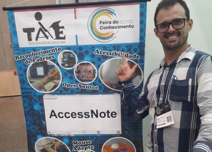 #descriçãodaimagem: foto do Junior. Ele é um homem branco de cabelos castanhos curtos. Ele tem barba e bigode, e usa óculos de grau. Junior está vestindo uma camisa xadrez. Ele está ao lado de um cartaz divulgando suas criações. Fim da descrição.