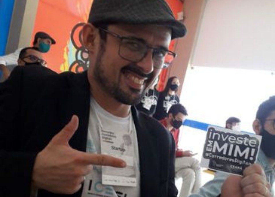 #descriçãodaimagem: foto do Junior. Ele é um homem branco de cabelos castanhos curtos. Ele tem barba e bigode, e usa óculos de grau. Junior está usando uma boina e veste uma camiseta branca e um terno preto. Ele está segurando uma pequena placa onde está escrito: investe em mim. Fim da descrição.