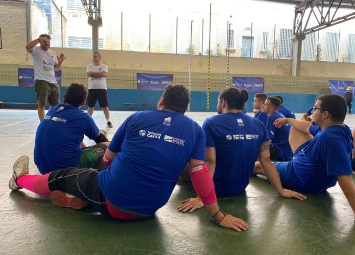 #descriçãodaimagem: Foto do treino de Futsal. Os atletas da CBDS estão sentados no chão da quadra e recebem instruções do técnico e do auxiliar técnico. Fim da descrição.
