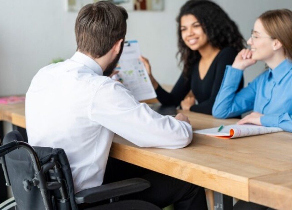 #descriçãodaimagem: foto de pessoas em uma reunião de trabalho. Uma das pessoas tem deficiência física. Fim da descrição.