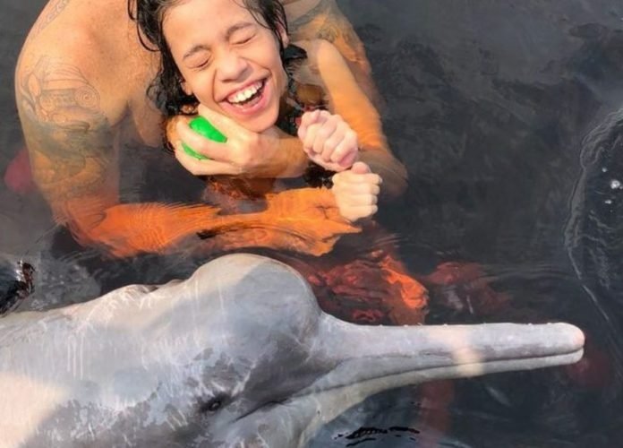 #descriçãodaimagem: Foto de uma menina interagindo com um boto nas águas do rio Negro, no Amazonas. A menina sorri. Ela está no colo de uma pessoa. Fim da descrição.