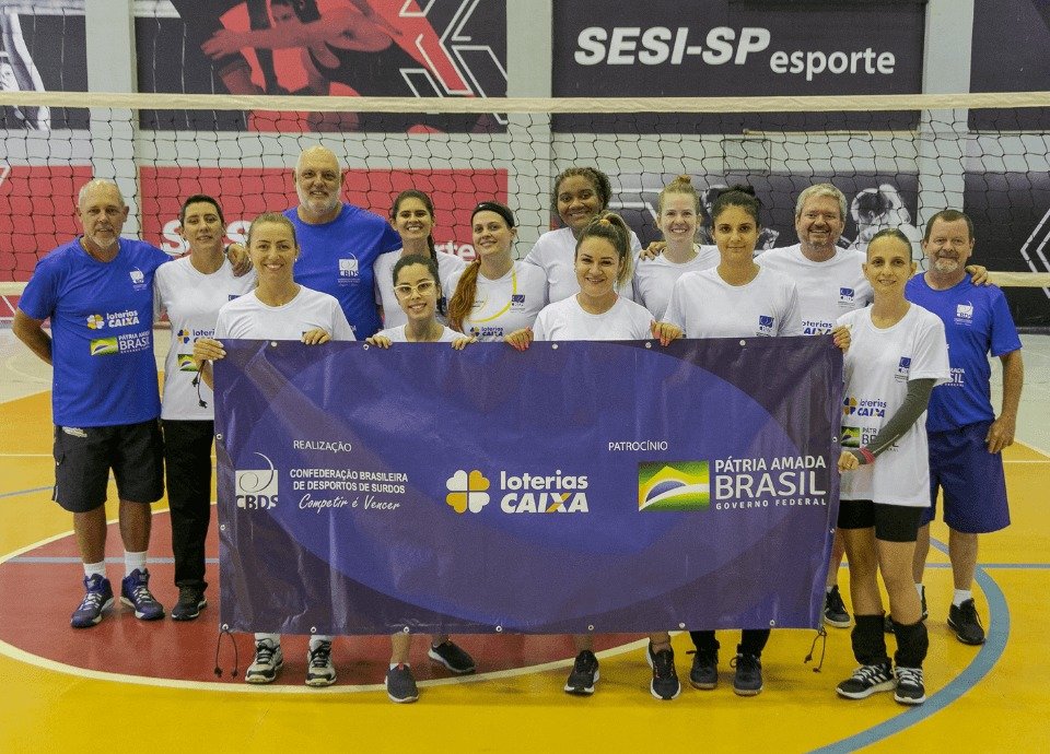 #descriçãodaimagem Foto das atletas surdas que participaram do treino seletivo de voleibol da CBDS. Elas estão na quadra onde o treino foi realizado e estão segurando uma faixa azul com os logos da CBDS e dos patrocinadores. Fim da descrição.