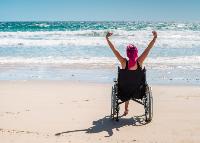 #descriçãodaimagem Foto de uma mulher na praia. A mulher tem cabelo cor de rosa e está em sua cadeira de rodas, de frente para o mar. Seus braços estão pra cima. Fim da descrição.