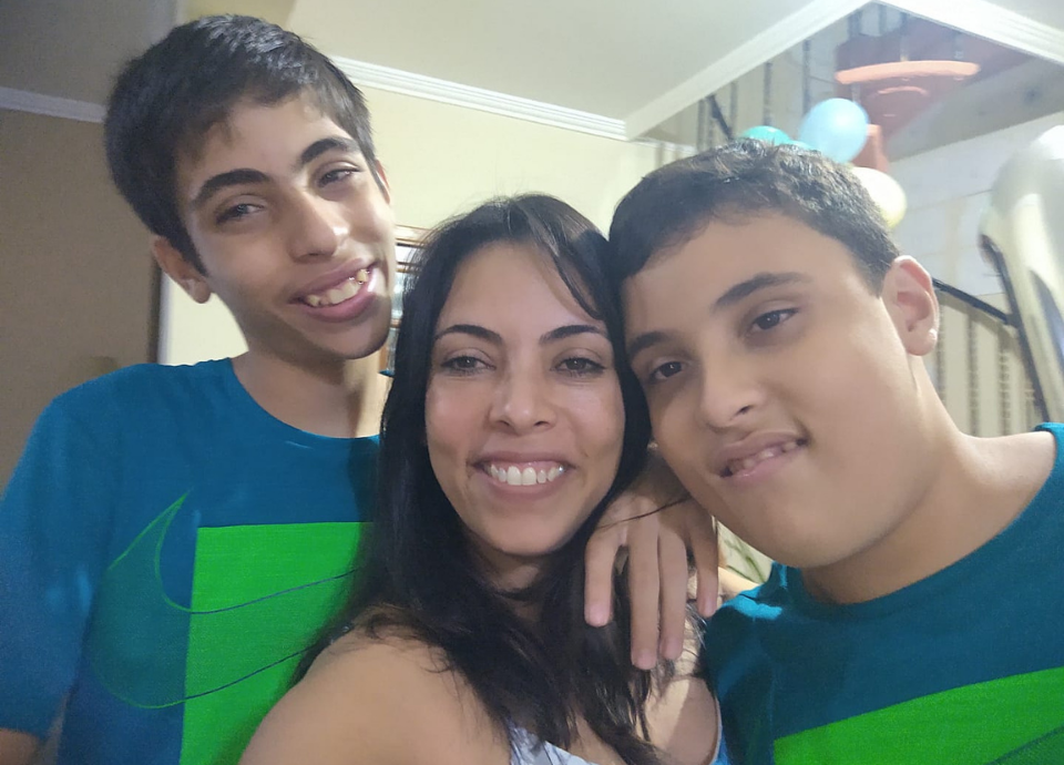 #descriçãodaimagem Foto de Fabiana com os filhos gêmeos. Ela é uma mulher branca de cabelos castanhos, compridos. Ela está entre seus dois filhos, que são gêmeos, mas não são identicos. Fim da descrição.