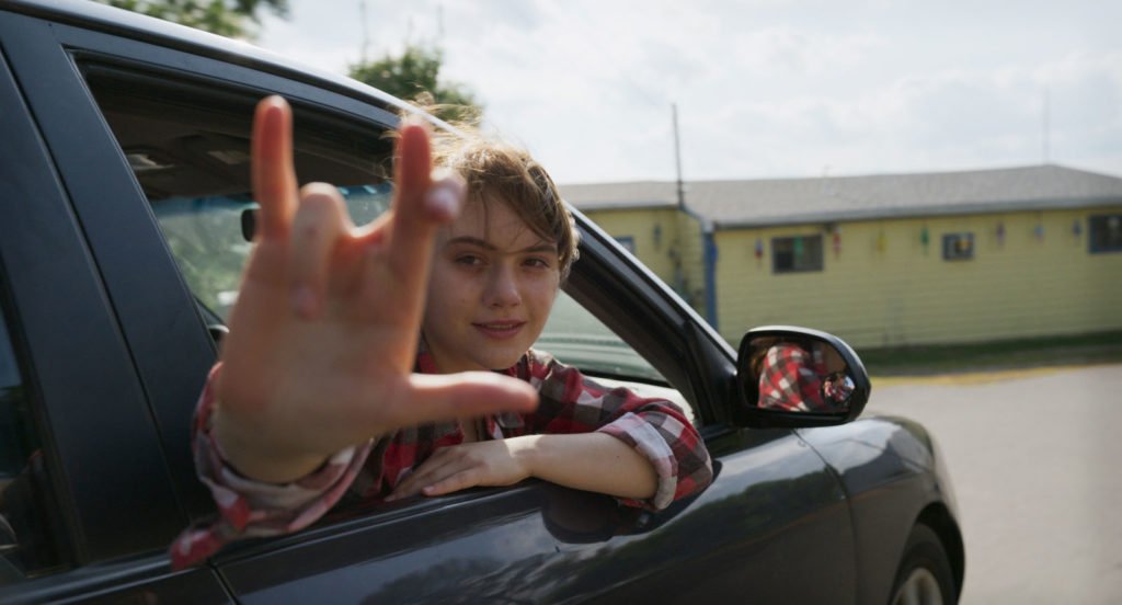 #descriçãodaimagem Foto de uma cena do filme No ritmo do coração. A protagonosta, uma jovem, está na janela de um carro e faz o sinal universal de Eu Amo Você, na língua de sinais. Fim da descrição.