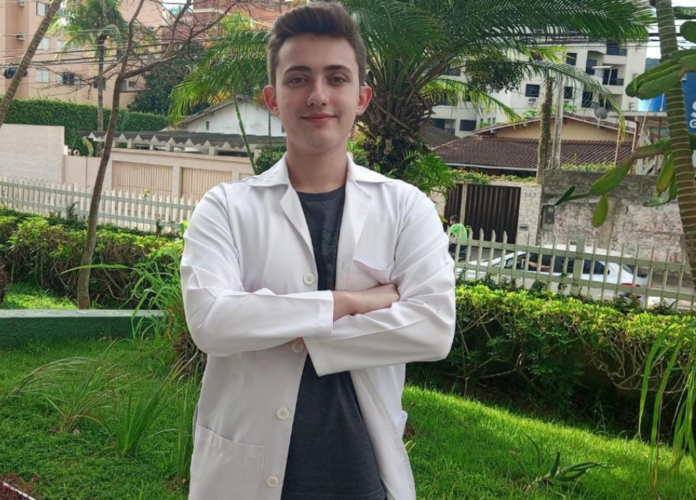#descriçãodaimagem Foto do Arthur. Ele é um jovem. Arthur está em um jardim. Ele é branco e tem cabelos castanhos curtos. Arthur está usando um jaleco branco e uma camiseta preta. Ele está com os braços cruzados. Fim da descrição.