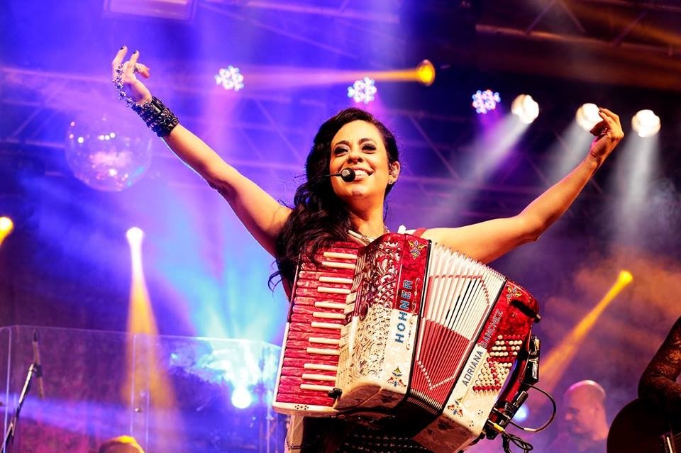 Descrição da imagem: Foto da sanfoneira Adriana Sanchez durante apresentação. Ela está no palco com sua sanfona. Adriana é uma mulher branca de cabelos castanhos londos. Ela está com os braços levantados e sorri. Fim da descrição.