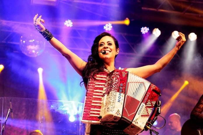 Descrição da imagem: Foto da sanfoneira Adriana Sanchez durante apresentação. Ela está no palco com sua sanfona. Adriana é uma mulher branca de cabelos castanhos londos. Ela está com os braços levantados e sorri. Fim da descrição.