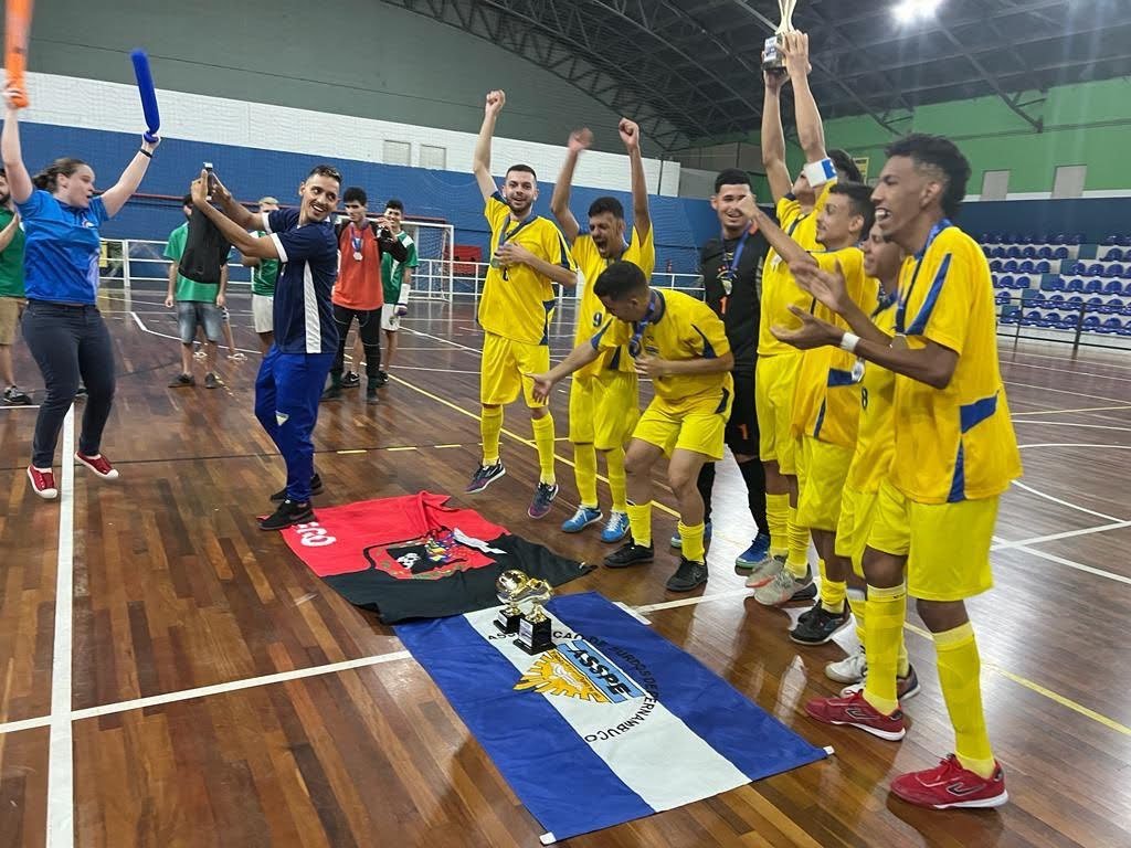 Descrição da imagem: Foto dos atletas da Associação de Surdos de Pernambuco comemorando o 1º lugar. Eles estão pulando na quadra. Fim da descrição.