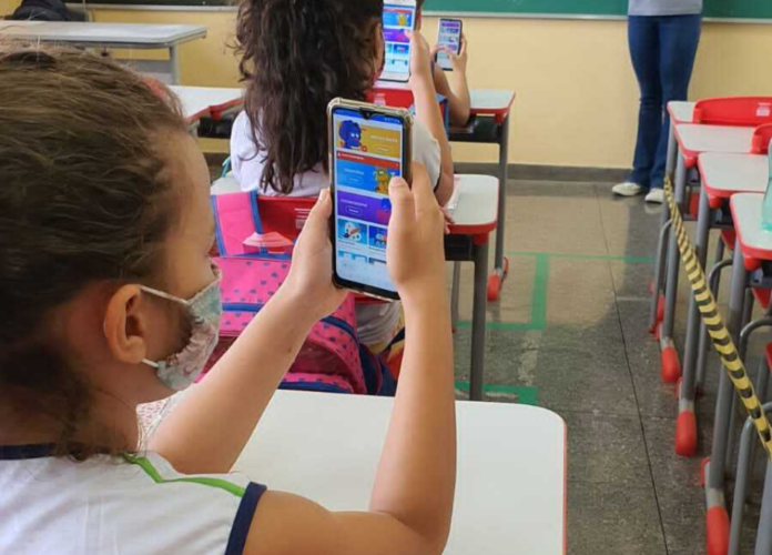 Descrição da imagem: Foto de uma menina na sala de aula. Ela está segurando um celular. Fim da descrição.