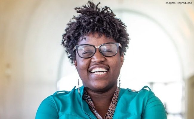 Descrição da imagem: Foto da cientista Nina da Hora. Ela é uma mullher negra. Nina tem cabelos castanhos, curtos. Ela está usando uma blusa verde e também óculos de grau. Nina está sorrindo. Fim da descrição.