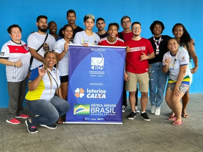 Descricão da imagem: Foto da equipe técnica e atletas da CBDS, entre outros participantes do torneio de Judô. Eles seguram um banner azul com o logo da CBDS e dos patrocinadores. Fim da descrição.