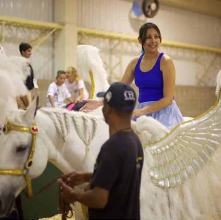 Descrição da imagem: Foto da equoterapeuta Eliane com o cavalo Raio. Raio é um cavalo branco e está com adornos. Fim da descrição.