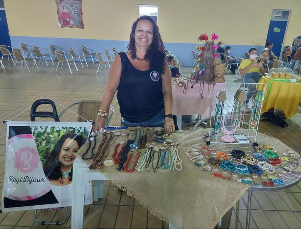 Descrição da imagem: Foto da empreendedora Helen, durante um evento. Ela está atrás de uma mesa, onde estão expostos seus trabalhos. Helen é uma mulher branca de cabelos castanhos cacheados. Fim da descrição.