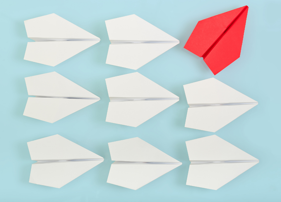Descrição da imagem: A imagem tem fundo azul claro. Nela estão nove aviões de papel. Oito deles são brancos e estão apontados para o lado esquerdo. Um deles é vermelho e está apontado para cima, na diagonal. Fim da descrição.