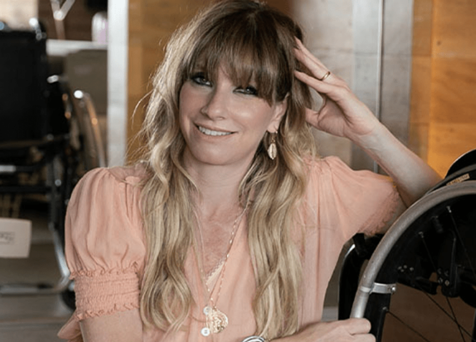 Descrição da imagem: Foto de Andrea Schwarz, conselheira da EqualWeb. Ela é uma mulher branca de cabelos loiros. Andrea está sentada ao lado de sua cadeira de rodas, e sorri. Ela está com uma blusa cor de rosa. Fim da descrição.