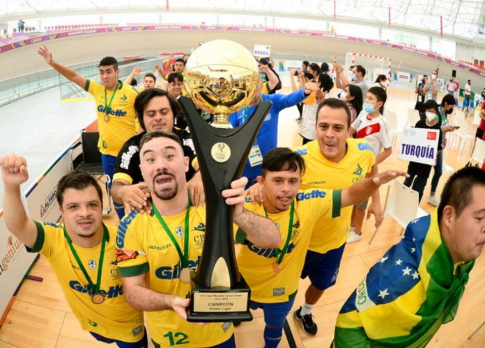 Descrição da imagem: Foto dos atletas da seleção brasileira de futsal Down. Eles estão comemorando e seguram o troféu. Fim da descrição.