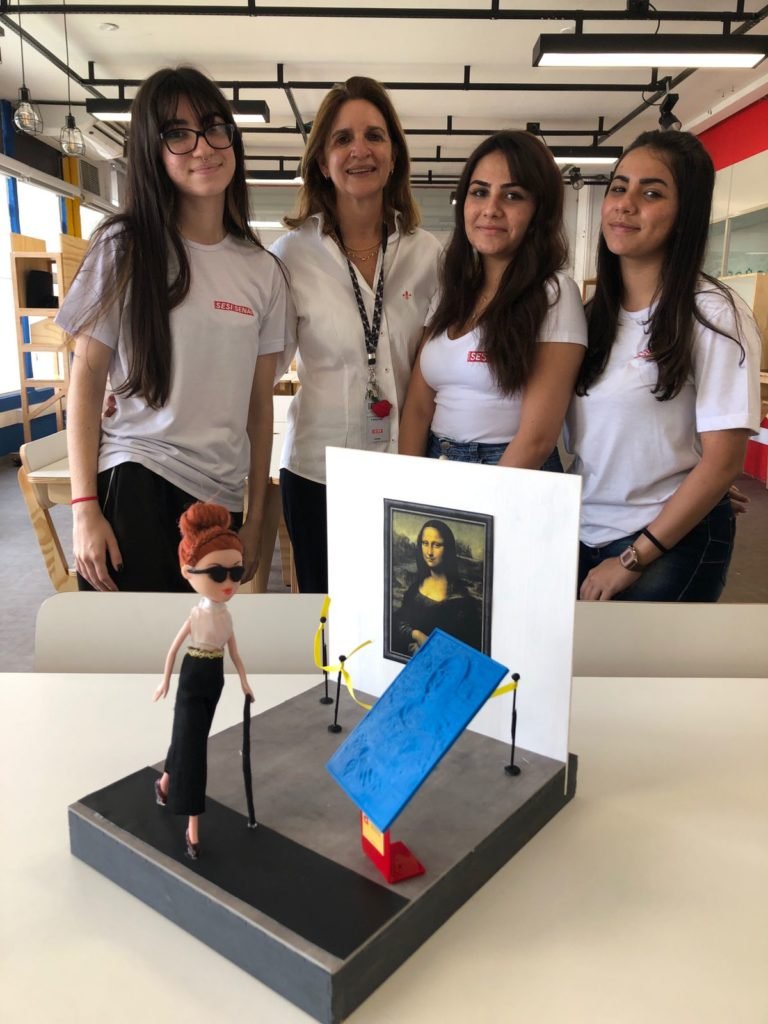 Descrição da imagem: Foto das três estudantes que idealizaram o projeto com a professora. Em primeiro plano está a obra com sua versão 3D. Fim da descrição.
