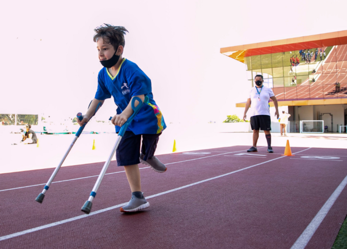 Descrição da imagem: Foto de uma atividade da Escola Paralímpica de Esportes do CPB. Um menino durante treino na pista de atletismo. O menino tem uma amputação da perna esquerda, abaixo do joelho. Ele usa muletas e está sendo observado pelo professor. Fim da descrição.