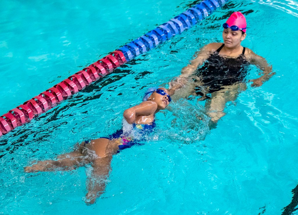 Descrição da imagem: Foto de uma atividade da Escola Paralímpica de Esportes do CPB. Uma menina durante treino de natação. A menina tem uma amputação nas duas pernas. Ela está nadando sob supervisão da professora. Fim da descrição.