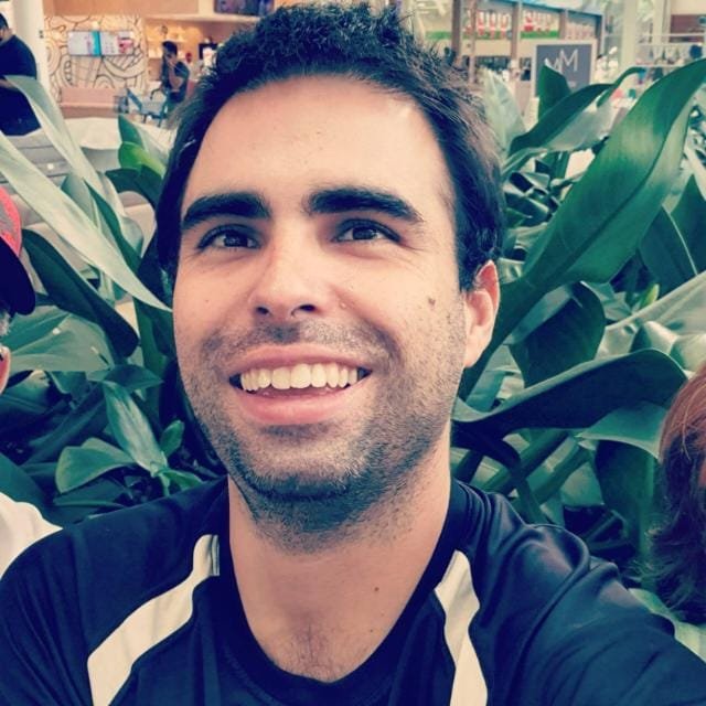 Descrição da Imagem: Foto de Sidney Zamboni, aluno da MUST University. Ele é um homem branco, tem cabelos escuros e sobrancelhas grossas. Na imagem, ele está sorridente. Usa uma camisa azul com figuras brancas. Ao seu fundo, aparece um jardim. Fim da descrição.