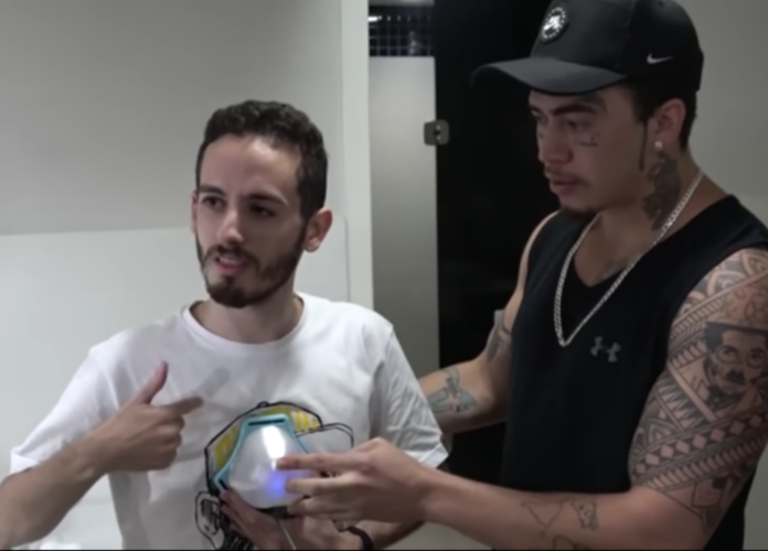 Descrição de imagem: foto do momento de teste da caixa de som. Whindersson Nunes está ao lado de um rapaz surdo. Ele segura o aparelho no peito do rapaz. Fim da descrição.
