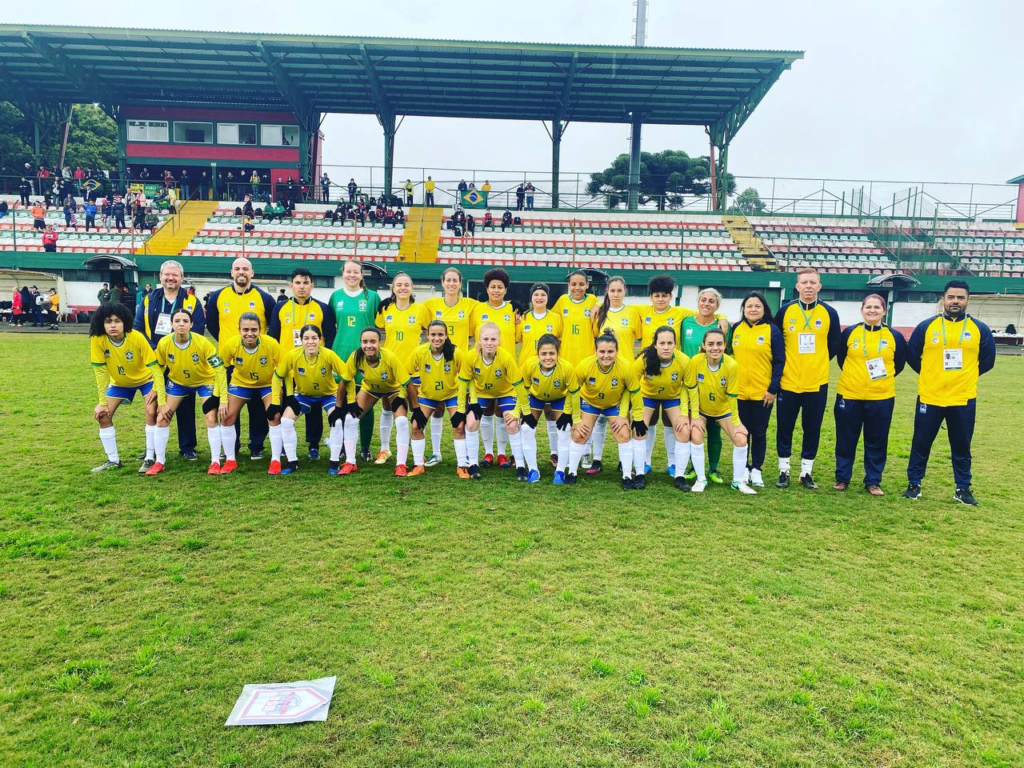 Descrição da imagem: foto das atletas da seleção brasileira de futebol feminino e membros da comissão técnica. Elas estão perfiladas no centro do campo, antes do jogo. As atletas usam uniforme amarelo, com excessão da goleira, que está com uniforme verde. Fim da descrição.