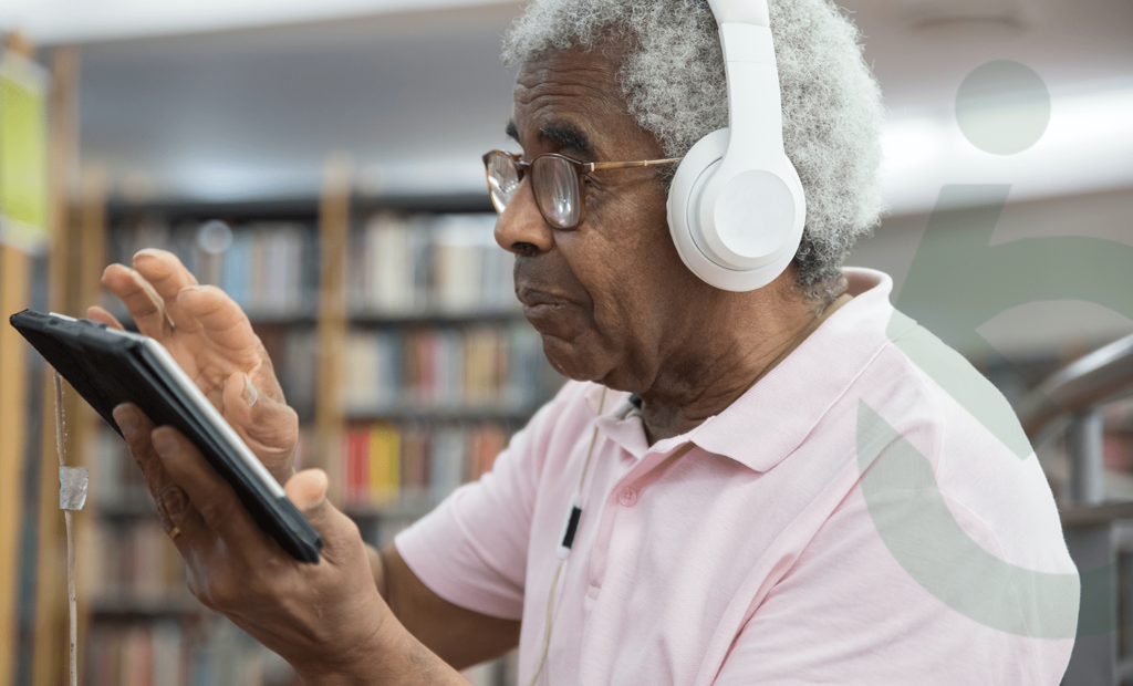 Descrição da imagem: foto de um idoso em uma livraria. Ele manuseia um tablet e está com fones de ouvido. Fim da descrição.