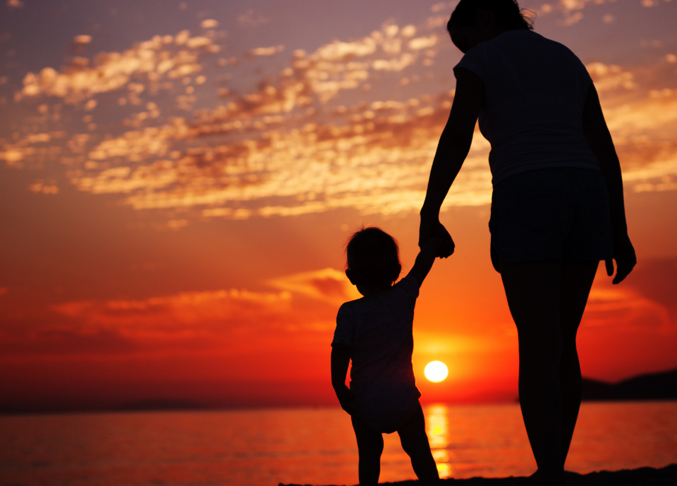 Descrição da imagem: foto da sombra de uma mulher com uma criança. Eles estão de mãos dadas e ao fundo vemos o pôr do sol. Fim da descrição.
