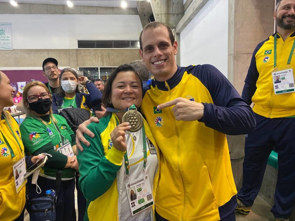 Descrição da imagem: foto da presidente da CBDS, Diana Kyosen, com o medalhista Guilherme Maia. Ele está segurando a medalha. Fim da descrição.