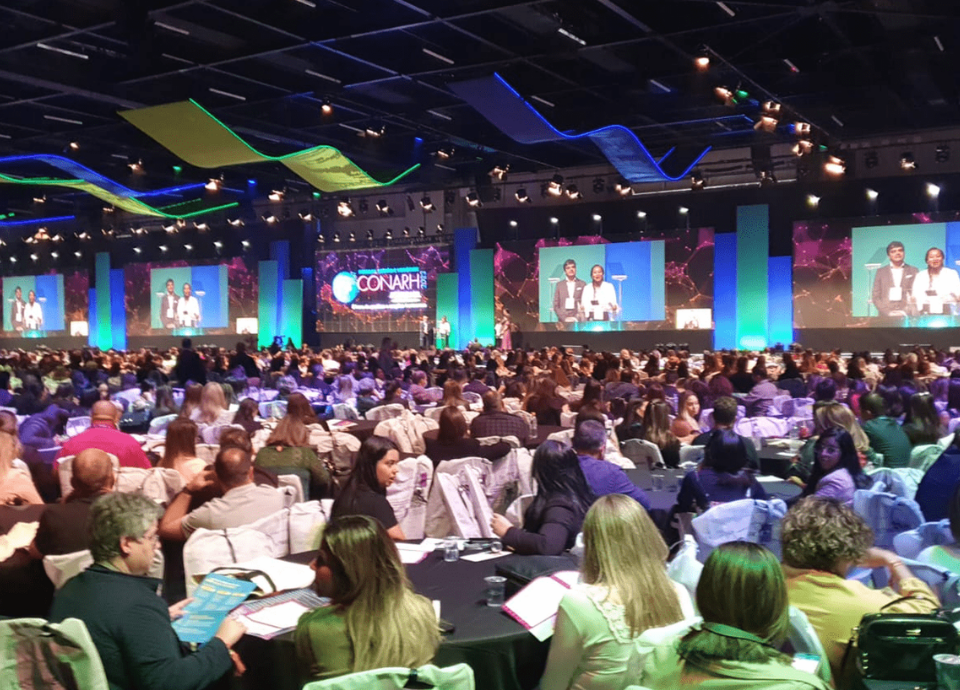 Descrição da imagem: foto do auditório do evento Conarh. Centenas de pessoas estão sentadas em um grande auditório. Ao fundo está um palco e grandes telões. Fim da descrição.