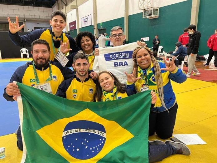 Descrição da imagem: foto do atleta Rômulo Crispim com a equipe de Judô. Eles estão com uma bandeira do Brasil. Fim da descrição.
