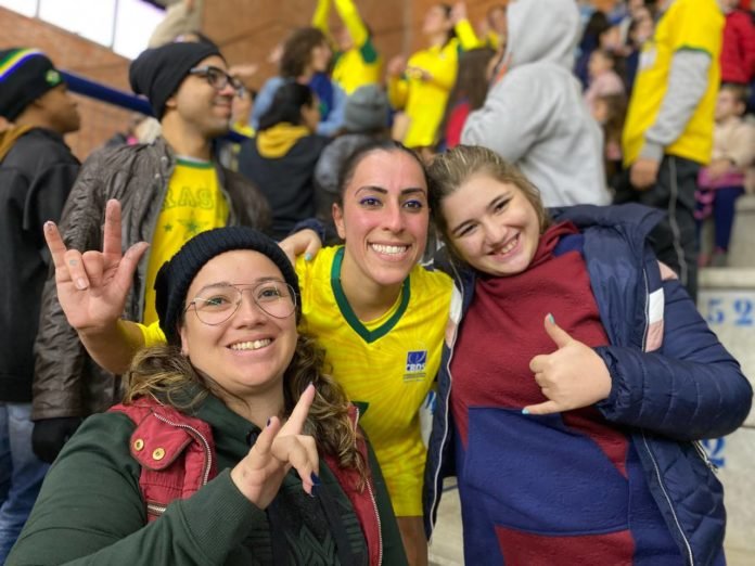 Descrição da imagem: foto de uma jogadora da seleção brasileira de handebol com duas torcedoras, na arquibancada do ginásio de esportes. As torcedoras fazem o sinal universal de 'eu te amo' na língua de sinais. Fim da descrição. partida de Handebol entre Brasil e Argentina. Fim da descrição.