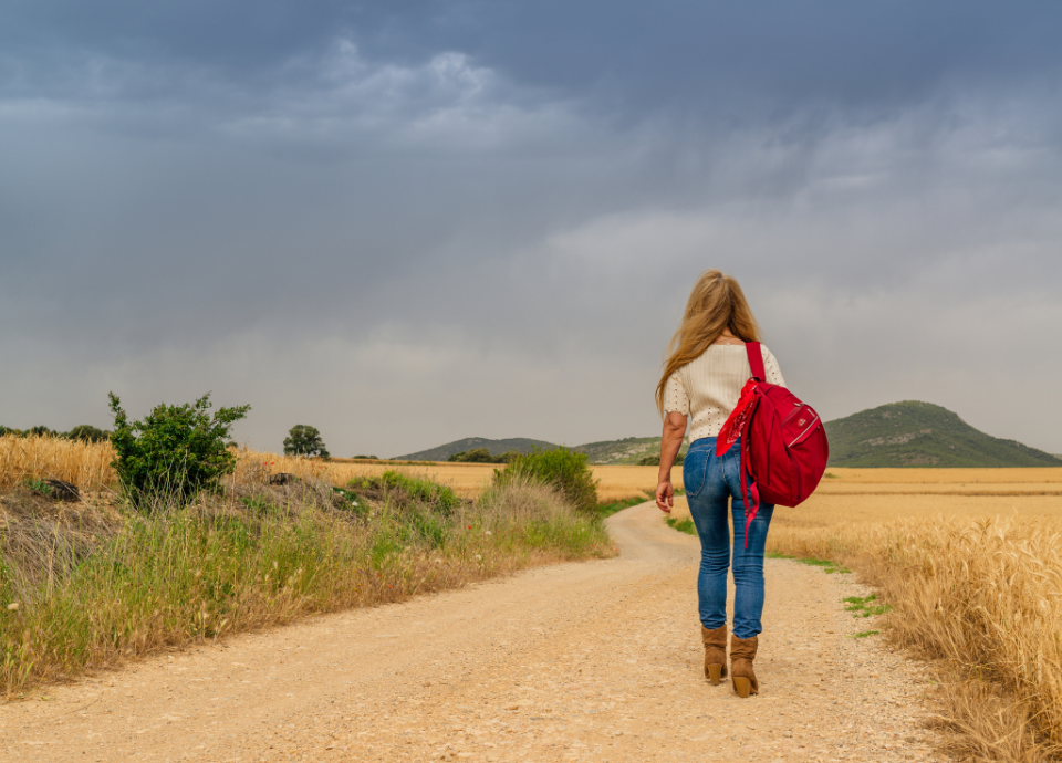 Descrição da imagem: foto de uma moça caminhando em uma estrada de terra. Ela usa blusa branca e calça jeans, e tem uma mochila vermelha nas costas. Fim da descrição.