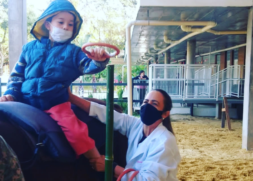 Descrição da imagem: foto de uma criança durante atendimento com profissional de equoterapia. Fim da descrição.
