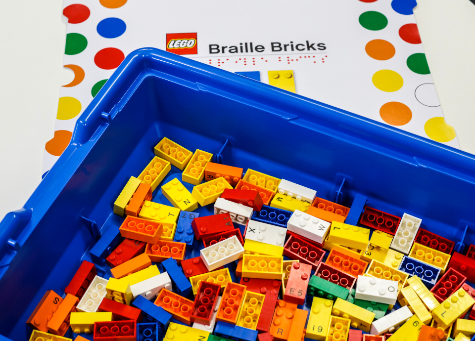 Descrição da imagem: foto da caixa do Braille Bricks. Ela é azul e está aberta. Dentro dela estão os blocos de montar coloridos. Na parte superior da imagem está o nome do brinquedo. Fim da descrição.