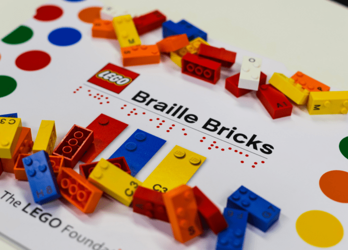 Descrição da imagem: foto de peças do Braille Bricks espalhadas sobre a embalagem. Fim da descrição.