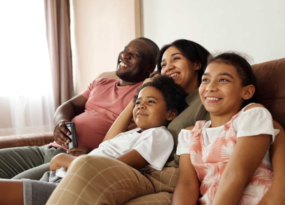 Descrição da imagem: foto de uma família, mãe, pai e duas crianças. Eles estão sentados no sofá e parecem se divertir assistindo TV. Fim da descrição.
