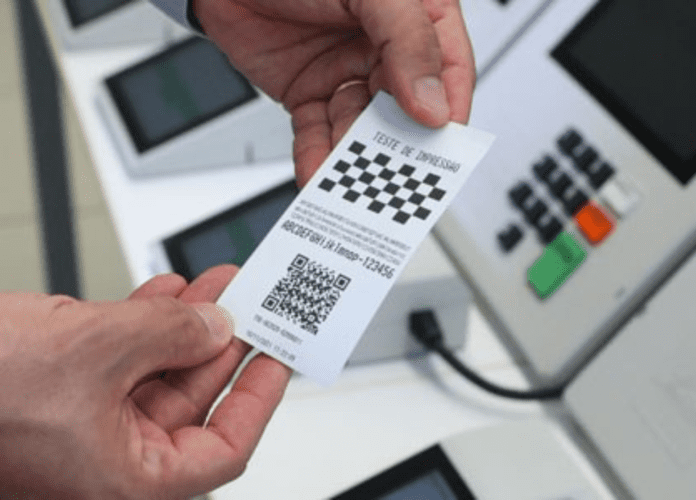 Descrição da imagem: foto de uma urna eletrônica. Em primeiro plano, duas mãos seguram um comprovante de votação. Fim da descrição.
