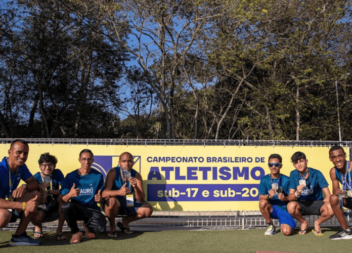 Descrição da imagem: foto dos alunos da Escola Paralímpica de Esportes do CPB. Eles estão posando com suas medalhas ao lado do banner de divulgação do Brasileiro de Atletismo sub-17 e sub-20. Fim da descrição.