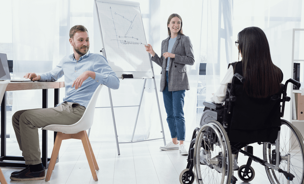 Descrição da imagem: foto de uma reunião de trabalho. Participam da reunião uma mulher, que está em pé ao lado de um quadro branco, um rapaz e uma mulher cadeirante. Fim da descrição.