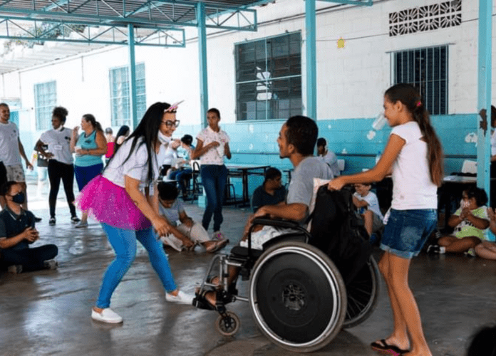 Descrição da imagem: foto de duas moças numa roda de capoeira no projeto de capoeira adaptada. Uma das moças é cadeirante. Fim da descrição.