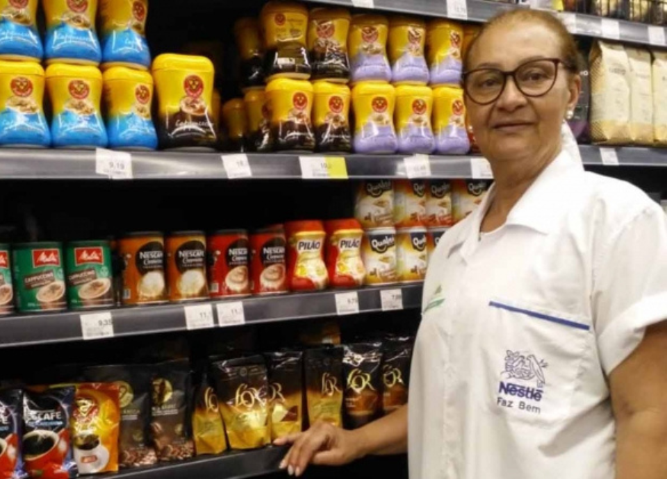 Descrição da imagem: foto de uma mulher idosa ao lado de uma prateleira de supermercado. Ela está usando um jaleco branco. Fim da descrição.