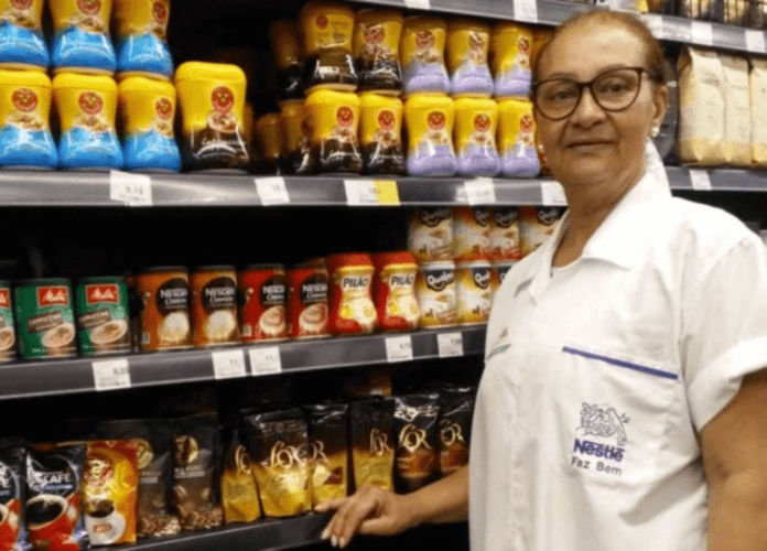 Descrição da imagem: foto de uma mulher idosa ao lado de uma prateleira de supermercado. Ela está usando um jaleco branco. Fim da descrição.