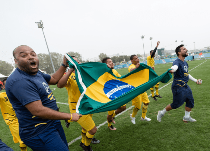 Descrição da imagem: foto dos atletas e técnicos da Olimpíadas Especiais Brasil comemorando em um campo de futebol. Eles estão segurando uma bandeira do Brasil. Fim da descrição.