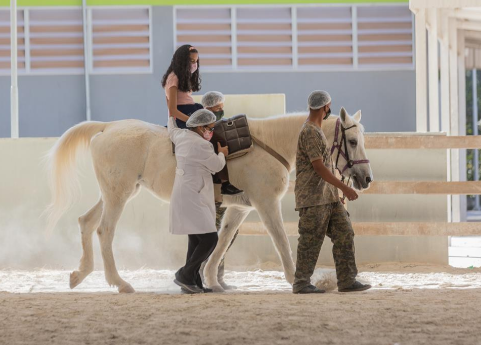 Descrição da imagem: foto do atendimento da equoterapia. Uma menina está montada em um cavalo branco. Um profissional guia o cavalo e duas profissionais realizam o atendimento na lateral do cavalo. Fim da descrição.